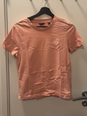 Persikofärgad t-shirt från Gant - Säljer en persikofärgad t-shirt från Gant med broderad logga på bröstet. T-shirten har rund halsringning och korta ärmar. Tillverkad i mjuk bomull för en skön känsla och enkel stil.