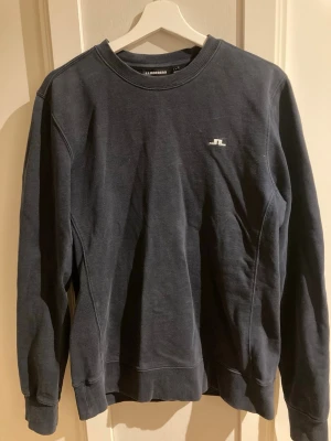 Mörkblå sweatshirt från J.Lindeberg - Mörkblå sweatshirt från J.Lindeberg med rund halsringning och diskret vitt logotryck på bröstet. Tröjan har långa ärmar och ribbade muddar vid ärmslut och nederkant. Perfekt för en avslappnad och stilren look.