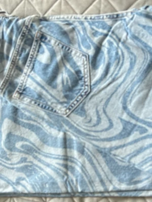 Ljusblå swirlmönstrade jeans - Unika ljusblå jeansbyxor med swirlmönster i vitt och blått. Klassisk femficksmodell i jeansmaterial med normal passform och raka ben. Perfekta för dig som vill sticka ut med ett coolt mönster och fräsch vibe.