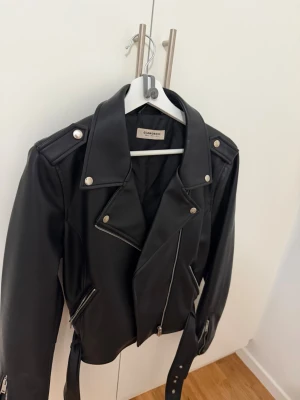 Svart  skinnjacka  - Snygg svart  skinnjacka från med silvriga detaljer, dragkedjor och bälte i midjan. Klassisk modell med bred krage och axelklaffar. Köpt från Nelly, använd en gång🩷