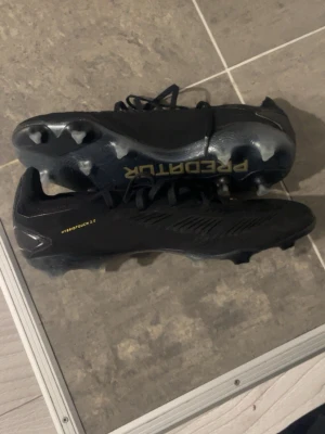 Adidas Predator svarta fotbollsskor - Svarta Adidas Predator fotbollsskor med gulddetaljer och snörning. Skorna har en låg profil och är tillverkade i syntetmaterial med texten 'HYBRIDTOUCH 2.0' på sidan. Perfekta för spel på gräsplaner.