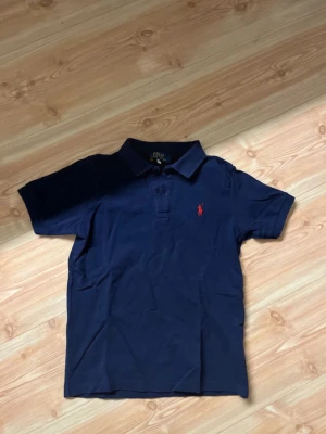 Marinblå pikétröja från Polo Ralph Lauren - Klassisk marinblå pikétröja från Polo Ralph Lauren med röd broderad logga på bröstet. Tröjan har korta ärmar, krage och knappar framtill. Perfekt för en stilren look.