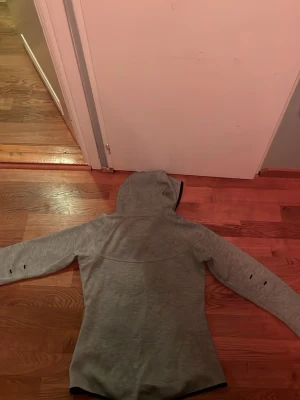 Ljusgrå hoodie med svarta detaljer - Snygg ljusgrå hoodie med dragkedja och svarta detaljer vid fickor och ärmslut. Utrustad med huva och tumhål på ärmarna för extra komfort. Perfekt för dig som gillar en sportig och avslappnad stil.