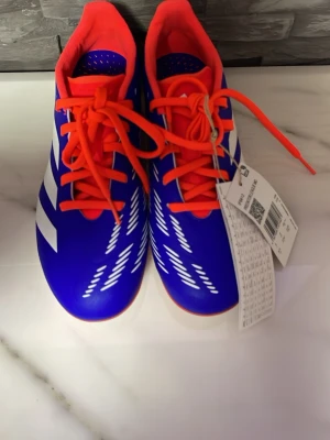 Blå-orange fotbollsskor från Adidas - Snygga fotbollsskor från Adidas i blått med vita detaljer och orange snörning samt sula. Skorna har klassisk snörning och mönstrad yttersula för bra grepp på planen. Helt nya med prislapp 