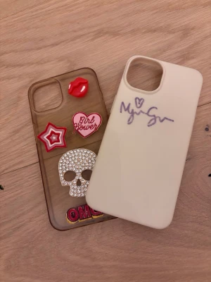 Två coola mobilskal med dekorationer - Säljer två unika mobilskal. Ett transparent skal med färgglada dekorationer som läppar, stjärna, hjärta, dödskalle med strass och texten 'OMG'. Det andra är ett beige skal med lila autograf av myragranberg Båda har öppning för kamera och passar modern smartphone. iPhone 13