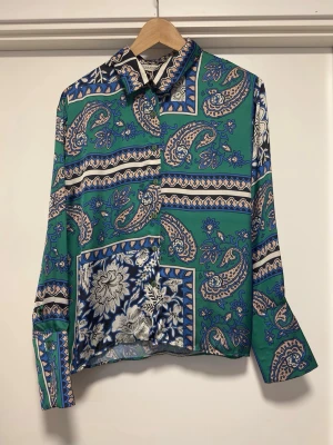 Grön paisleymönstrad skjorta - Snygg skjorta med paisley- och blommönster i grönt, blått, vitt och beige. Skjortan har klassisk krage, knappar framtill och långa ärmar. Materialet känns lätt och svalt, perfekt för dig som gillar färgstarka och unika plagg.