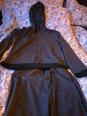 Mörkgrått loungewear-set med huva - Mörkgrått loungewear-set i mjukt material med matchande tröja och byxor. Tröjan har huva, knappar vid halsen och svarta muddar. Byxorna har resår i midjan och muddar vid bensluten för extra komfort. Perfekt för chill hemma.