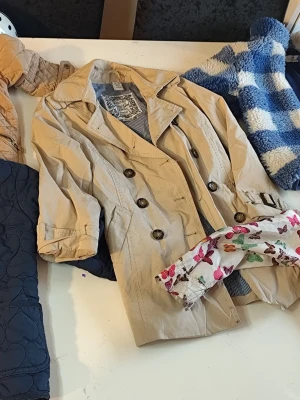 Beige trenchcoat med knappar stl 110/116 - Stilren beige trenchcoat med dubbelknäppning och klassisk krage. Jackan har långa ärmar som kan vikas upp och dekorativa knappar framtill. Perfekt för dig som gillar tidlös och enkel stil.Du är varmt välkommen att kika på mina andra annonser.