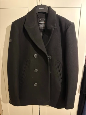 Svart ullkappa från Superdry - Stilren svart skepparkavaj från Superdry Merchant Line med dubbelknäppning och bred krage. Jackan är tillverkad i ullblandning och har snedställda fickor framtill. Perfekt för dig som gillar klassisk marin stil.