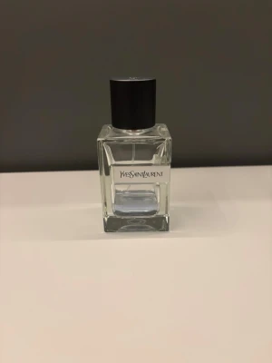 Yves Saint Laurent Y EdT parfym - Fräsch parfym från Yves Saint Laurent i en stilren, genomskinlig glasflaska med svart lock och en stor silverfärgad Y-detalj på sidan. Doften är lätt blåtonad och flaskan har en modern, kantig design. Ungefär 10ml kvar utav 60ml flaskan.