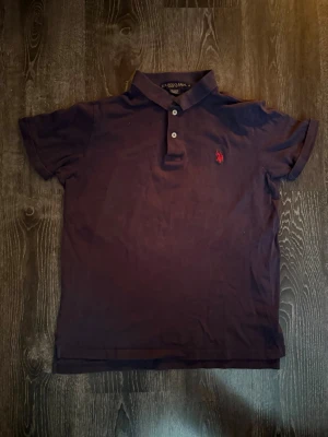 Mörkblå pikétröja från U.S. Polo Assn. - Klassisk mörkblå pikétröja från U.S. Polo Assn. med korta ärmar och två knappar framtill. Tröjan har en röd broderad logga på bröstet och är tillverkad i bomull för en skön känsla. Perfekt till jeans eller shorts.