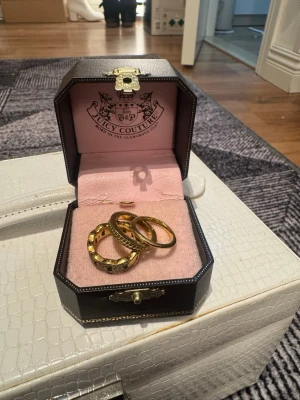 Guldfärgade ringar Juicy Couture - Säljer ett set med guldfärgade ringar från Juicy Couture. Ringarna har olika mönster, varav en är dekorerad med gnistrande stenar och en har hamrad yta. Kommer i en snygg svart ask med rosa insida och Juicy Couture-logga.