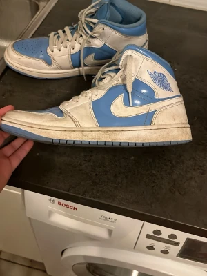 Nike Air Jordan 1 blå och vita - Nike Air Jordan 1 sneakers i blått och vitt med klassisk hög siluett. Skorna har snörning, rund tå och är tillverkade i skinn med perforerade detaljer på tån. Swoosh-logga på sidan och Air Jordan-logga vid ankeln. Perfekta för dig som gillar streetwear.