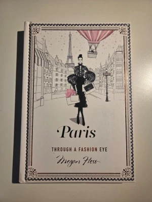 Paris Through a Fashion Eye - Stilren och elegant bok med hård pärm, illustrerad framsida i vitt, svart och rosa med motiv av Paris och mode. Bokens kanter har en lyxig kopparfärgad finish. Perfekt för dig som älskar mode, Paris och snygg design.