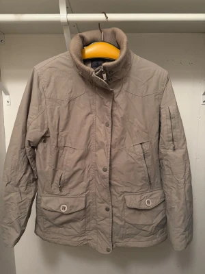 Beige vindjacka med hög krage - Stilren beige vindjacka med hög ribbad krage och dragkedja framtill. Jackan har flera fickor med både dragkedja och knappar, samt diskreta sömdetaljer. Perfekt för kyliga dagar och enkel att matcha med olika outfits.