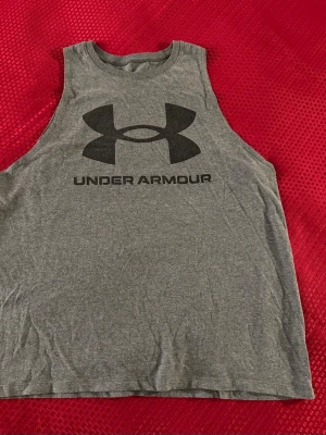 Grått linne från Under Armour - Snyggt grått träningslinne från Under Armour med stort svart logotryck på bröstet. Perfekt för gymmet eller löprundan. Mjukt och lätt material som andas bra.