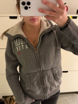 Fodrad grå zip up hoodie  Hollister - Mysig grå hoodie från Hollister med vit teddyfoder på insidan, dragkedja framtill och två stora fickor. Tryck på bröstet och justerbar huva med snören. Perfekt för chill dagar och enkel att matcha med jeans eller leggings.