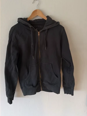 Svart zip hoodie med fickor - En svart hoodie med dragkedja framtill och två stora magfickor. Tröjan har huva med dragsnören och ribbade muddar vid ärmar och nederkant. Perfekt för en avslappnad och chill stil. Materialet känns mjukt och bekvämt, troligen bomull eller bomullsblandning.
