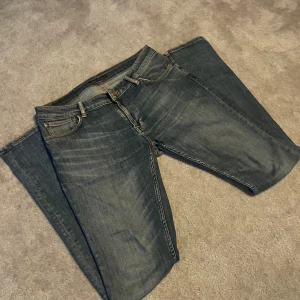 Nudie Jeans - Säljer ett par mörkblå jeans från Nudie Jeans med klassisk femficksdesign och snygga slitningar. Jeansen har raka ben och subtila kontrastsömmar samt Nudie-loggan på bakfickan. Perfekta för dig som gillar en stilren och avslappnad look.