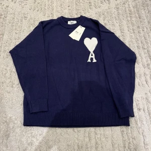 Mörkblå stickad tröja med hjärtdetalj - Mörkblå stickad tröja från AMI Paris med vit hjärtformad detalj och bokstaven A på bröstet. Tröjan har rund halsringning, långa ärmar och ribbstickade muddar vid ärmslut och nederkant. Perfekt för en trendig och avslappnad look.