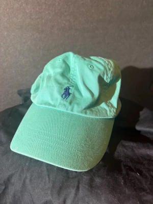 Mintgrön keps från Polo Ralph Lauren - Snygg mintgrön keps från Polo Ralph Lauren med klassisk böjd skärm och mörkblå broderad logga framtill. Tillverkad i bomull och har justerbar rem bak för perfekt passform. Perfekt accessoar för en avslappnad och trendig stil.