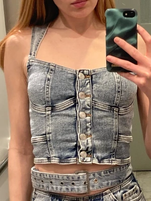 Denim korsettopp korsett topp crop bustier jeanstyg  - Snygg croppad denim korsettopp från Amisu med knappar framtill och breda axelband. Toppen har en cool tvättad blå färg och markerade sömmar som ger en edgy look. Perfekt att styla med jeans eller kjol för en trendig outfit.