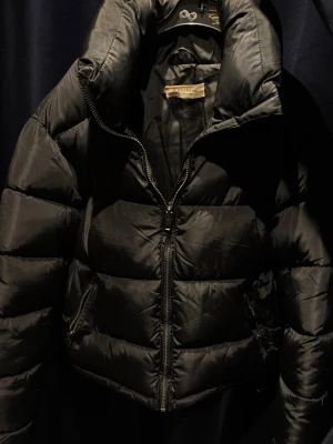 Svart pufferjacka - Säljer en svart pufferjacka från Glamorous med hög krage och quiltad design. Jackan har dragkedja framtill och två sidofickor med dragkedja. Materialet är syntet och polyester, perfekt för kalla dagar. Jackan har en loose passform och långa ärmar.