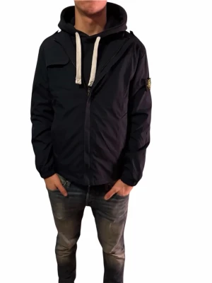 Mörkblå vindjacka från Stone Island - Säljer en mörkblå vindjacka från Stone Island utan huva. Jackan har en klassisk patch på ärmen och är tillverkad i polyester. Snygg och stilren modell som passar perfekt till streetwear. Fodret har coolt medaljmönster.