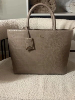 Beige handväska i krokopräglat skinn - Stilren beige handväska från Don Donna i krokopräglat skinnimitation. Väskan har två handtag och en fyrkantig form, perfekt för att bära dina viktigaste saker. Snygg detalj med hängande tag och diskret logga framtill.