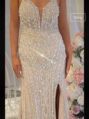 Glittrig långklänning med slits + släp - Elegant långklänning i ljus champagnefärg med glittriga paljettdetaljer och djup v-ringning. Klänningen har smala axelband, figurnära passform och en hög slits framtill. Perfekt för bal eller festliga tillfällen. Lägg bud, pris kan diskuteras vid snabb affär. 