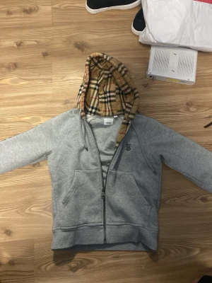 Grå zip hoodie från Burberry - Säljer en grå hoodie från Burberry med dragkedja och klassiskt rutigt foder i huvan. Hoodien har fickor framtill och en diskret Burberry-logga på bröstet. Perfekt för dig som gillar stilrena plagg med en twist. Materialet är mjuk bomullsmix.