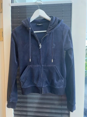 Mörkblå velourhoodie Juicy Couture - Mjuk och lyxig mörkblå velourhoodie från Juicy Couture med dragkedja, huva och fickor framtill. Har snygga detaljer som JC-logga på bröstet och metall-dragkedja med J-hänge. Perfekt för en chill och trendig look.