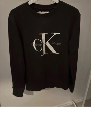 Svart sweatshirt från Calvin Klein - Svart sweatshirt från Calvin Klein Jeans med stort vitt CK-tryck på bröstet. Klassisk rund halsringning och ribbade muddar vid ärmslut och nederkant. Perfekt för en stilren och avslappnad look.