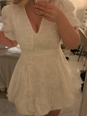 Vit kortklänning med puffärm - Supersöt vit kortklänning med puffärmar och v-ringning. Klänningen har ett blommigt mönster i tyget och markerad midja som ger en feminin siluett. Perfekt för studenten!🩷 Ny med prislappen kvar