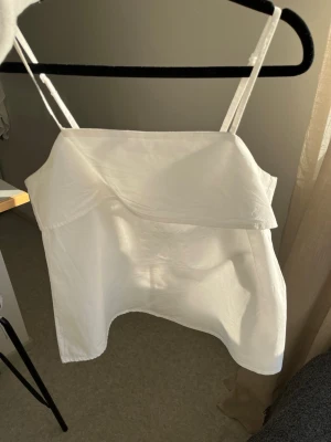 Vit topp - Snygg vit axelbandslös linnetopp med tunna axelband och en bred volangdetalj över bysten. Toppen har en rak passform och dragkedja i ryggen för enkel på- och avtagning. Perfekt för varma dagar och enkel att styla till olika looks.