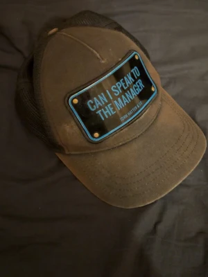 Svart keps med blå textplatta - Cool svart keps med böjd skärm och mesh på sidorna. Framtill sitter en svart och blå platta med texten 'CAN I SPEAK TO THE MANAGER?' i blått. Kepsen har en modern streetstyle-vibe och passar perfekt till en avslappnad look. Org saker följer med!