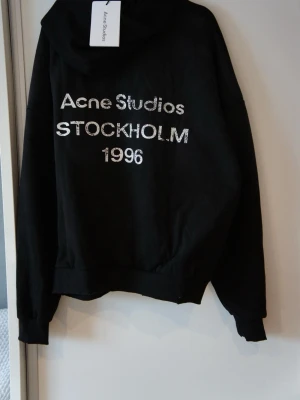 Svart hoodie Acne Studios Stockholm - Svart hoodie från Acne Studios med tryck i vitt framtill där det står 'Acne Studios STOCKHOLM 1996'. Oversized passform med huva och ribbade muddar. Perfekt för en avslappnad och trendig look. ❗️OBS❗️ - Stor i storlek, skriv till mig innan köp! 🙌🏼