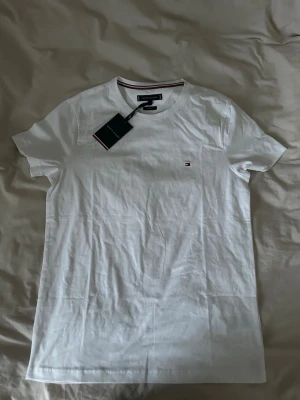 Vit t-shirt från Tommy Hilfiger - Klassisk vit t-shirt från Tommy Hilfiger med rund halsringning och diskret logga broderad på bröstet. Tillverkad i mjuk bomull och har korta ärmar. Perfekt basplagg med stilrena detaljer och en liten etikett i nacken.