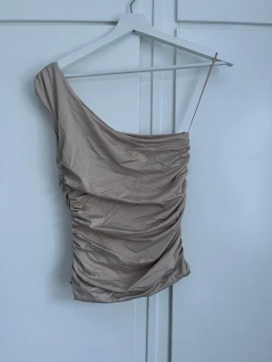 Beige oneshoulder topp - Trendig beige oneshoulder topp med draperad effekt och smal axelrem på ena sidan. Snyggt åtsittande passform och tillverkad i ett mjukt syntetmaterial. Perfekt för dig som vill sticka ut med en unik topp.