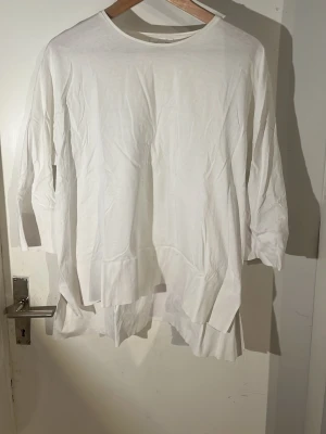 Vit oversized topp med trekvartsärm - En enkel och stilren vit topp med rund halsringning och trekvartsärm. Modellen är oversized med slitsar i sidorna och rak passform. Perfekt för en avslappnad look och lätt att matcha med olika outfits.