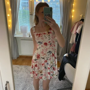 Blommig sommarklänning från Jacqueline de Yong - Söt vit klänning med blommigt mönster i rött, rosa och blått från Jacqueline de Yong. Klänningen har breda axelband, markerad midja och lätt utställd kjol. Perfekt för varma dagar och har en luftig känsla.