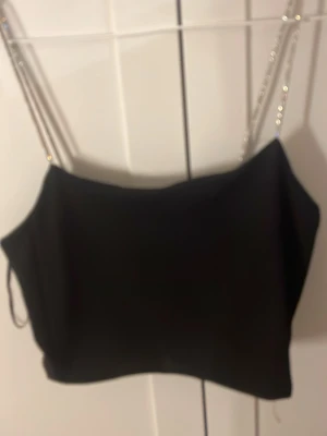 Svart croppad linnetopp med glitterband - Snygg svart croppad linnetopp med tunna glitterband som glittrar som axelband. Enkel och stilren design som passar perfekt till många olika outfits. Materialet känns mjukt och följsamt mot huden. Storlek L men är liten i storlek 
