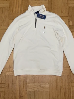 Beige halvzip från Polo Ralph Lauren - Klassisk vit halvzip-tröja från Polo Ralph Lauren med broderad logga på bröstet. Tröjan har hög krage, dragkedja och ribbade muddar. Tillverkad i mjukt bomullsmaterial som känns skönt mot huden. Perfekt för en clean och stilren look.