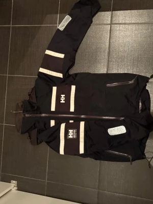 Mörkblå Helly Hansen vindjacka - Svart vindjacka från Helly Hansen med vita reflexdetaljer och HH-logga på bröstet och huvan. Jackan har dragkedja framtill, hög krage och flera praktiska fickor. Tillverkad i slitstark polyester och syntetmaterial, perfekt för blåsiga dagar.