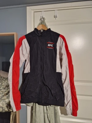 UFC vindjacka i svart, rött och vitt - Sportig vindjacka från UFC med svart bas, vita paneler och röda detaljer på ärmarna. Jackan har hög krage, dragkedja framtill och två sidofickor. Perfekt för dig som gillar streetwear och sportig stil.