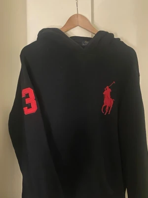 Svart hoodie från Polo Ralph Lauren - Svart hoodie med stor röd Polo-logga på bröstet och röd siffra 3 på ärmen. Klassisk huva och avslappnad passform, perfekt för en sportig och stilren look.