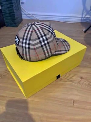 Burberry keps - Snygg keps från Burberry i klassiskt beige rutigt mönster med svarta, vita och röda linjer. Kepsen har böjd skärm och justerbar rem baktill. Perfekt accessoar för en trendig och stilren look.