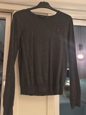 Mörkgrå stickad tröja från Ralph Lauren - Mörkgrå tunn stickad tröja från Ralph Lauren med rund halsringning och diskret röd logga broderad på bröstet. Tröjan har lång ärm och ribbade muddar vid ärmslut och nederkant. Perfekt till en enkel och stilren look.
