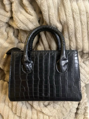 Svart handväska i krokopräglat skinn - Stilren svart handväska med krokopräglat mönster och två rejäla handtag. Väskan har en boxig form och är tillverkad i skinnimitation med struktur som påminner om krokodil. Perfekt accessoar för att lyfta din outfit.