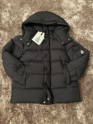 Svart Moncler dunjacka med huva - Snygg svart dunjacka från Moncler med klassisk pufferdesign och stor huva. Jackan har dragkedja och knappar framtill, två sidofickor och Moncler-logga på ärmen. Perfekt för kalla dagar och riktigt trendig look.
